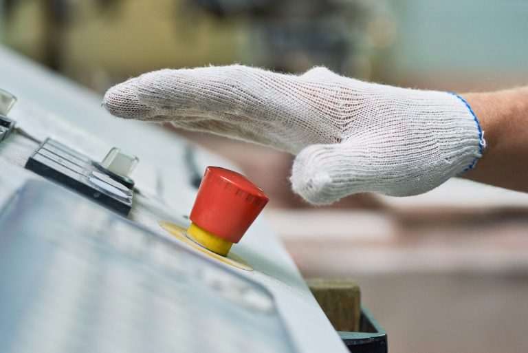 Not-Halt-Taster an einer Industriemaschine Hand mit weißem Schutzhandschuh über einem Not-Halt-Taster an einem Maschinenbedienpult